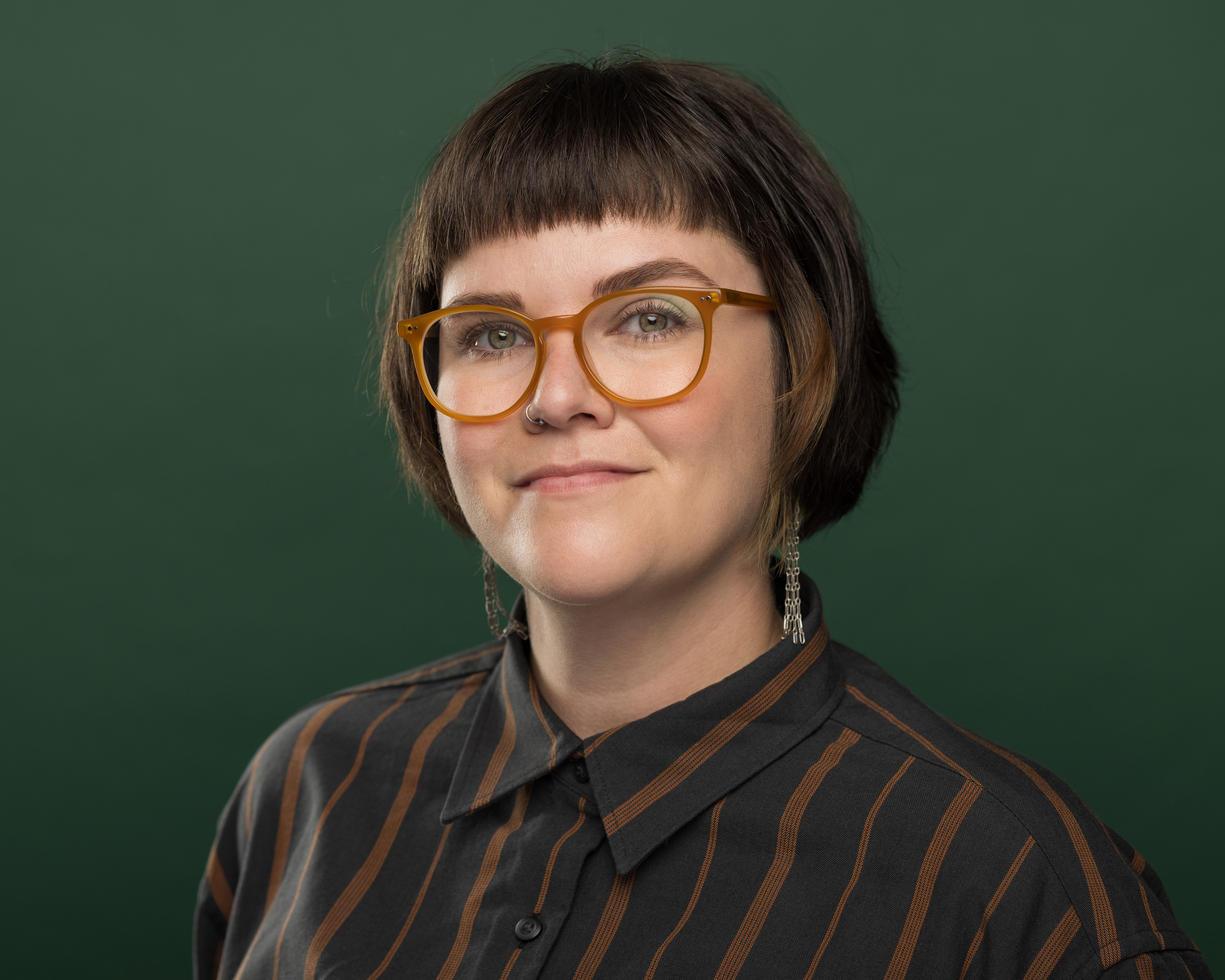 A woman with short brown hair, looking directly at the camera, in front of a dark green backdrop. She has orange plastic glasses and is wearing a dark blue shirt with tan strips.